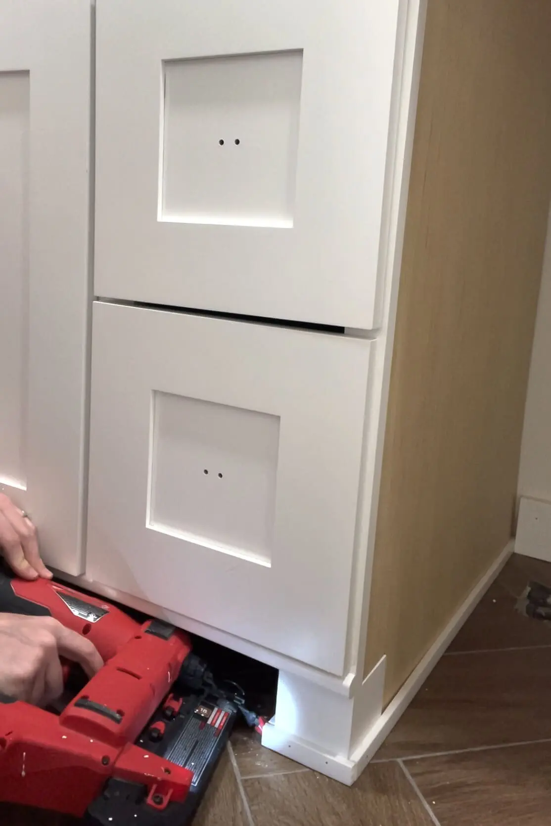 Close-up of the base of a white shaker cabinet with a person using a red nail gun to install white baseboard trim during a bathroom renovation project.